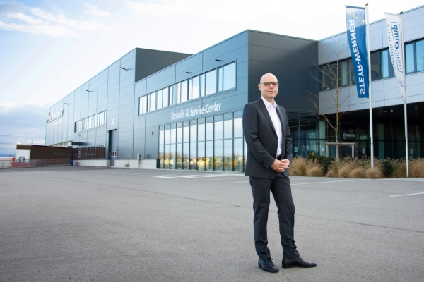 Ein Mann im Anzug steht auf einer leeren Straße vor einem modernen Industriegebäude - DEXIS Austria Zentrale in Pasching.. Die Folierung „Technik & Service-Center“ glänzt auf der Gebäudefront. Zwei hohe Fahnen flattern vor dem bewölkten Himmel.